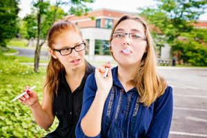 Teenage girl teaching an unsure friend how to smoke using an e-cigarette. 